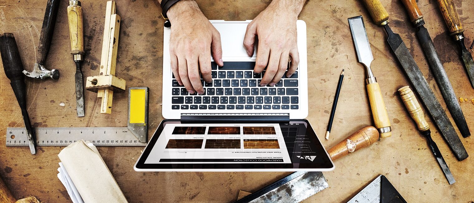 Ein Laptop steht auf einer Werkbank in einer Werkstatt. Daran sitzt ein Handwerker und schreibt etwas.