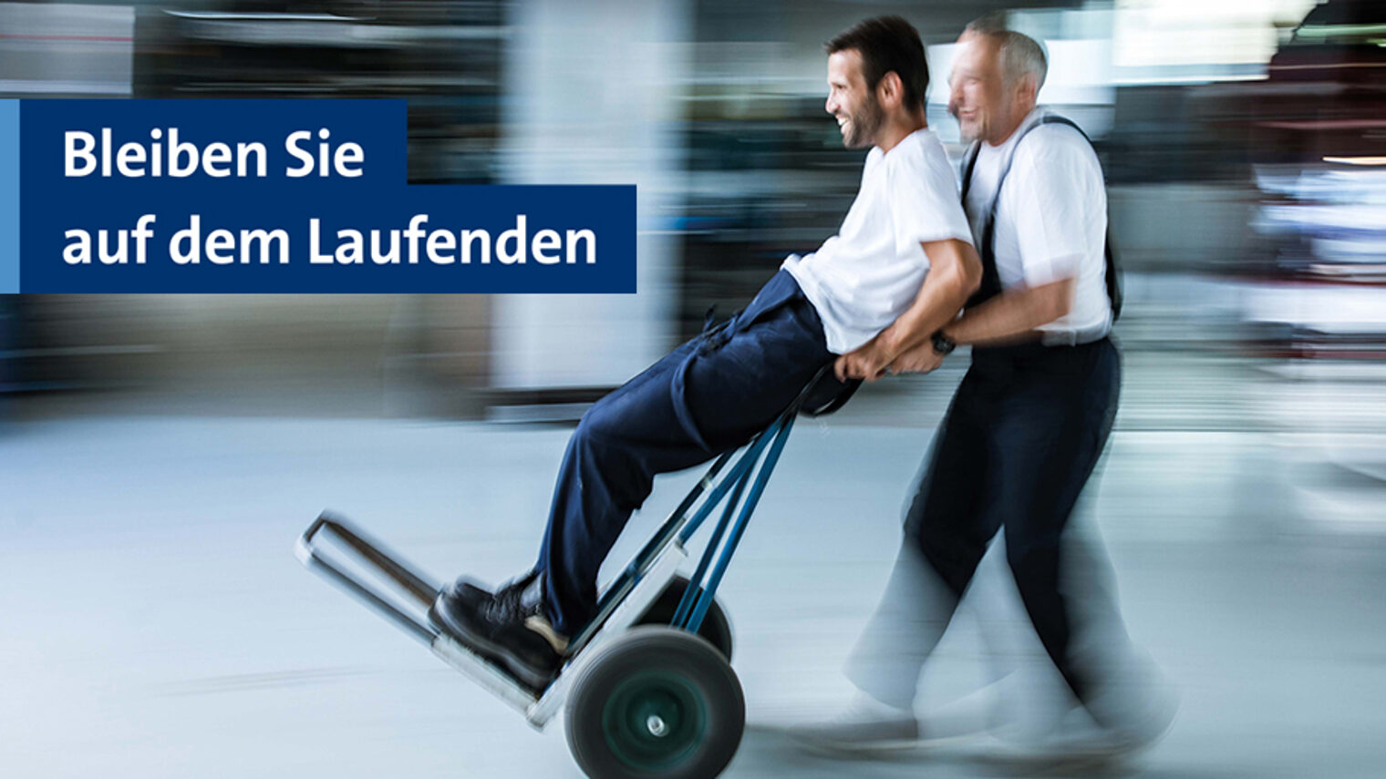 Zwei Handwerker, die auf einer Sackkarre durch eine Werkstatt fahren. Bleiben Sie auf dem Laufenden mit dem Newsletter der Handwerkskammer Berlin.