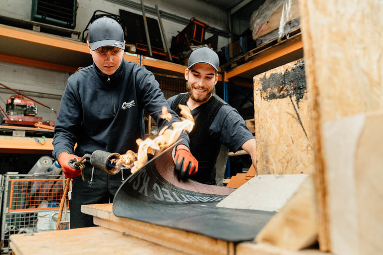 Zwei Dachdecker-Azubis beim Arbeiten in der Werkstatt, der eine hält eine Fackel in der Hand