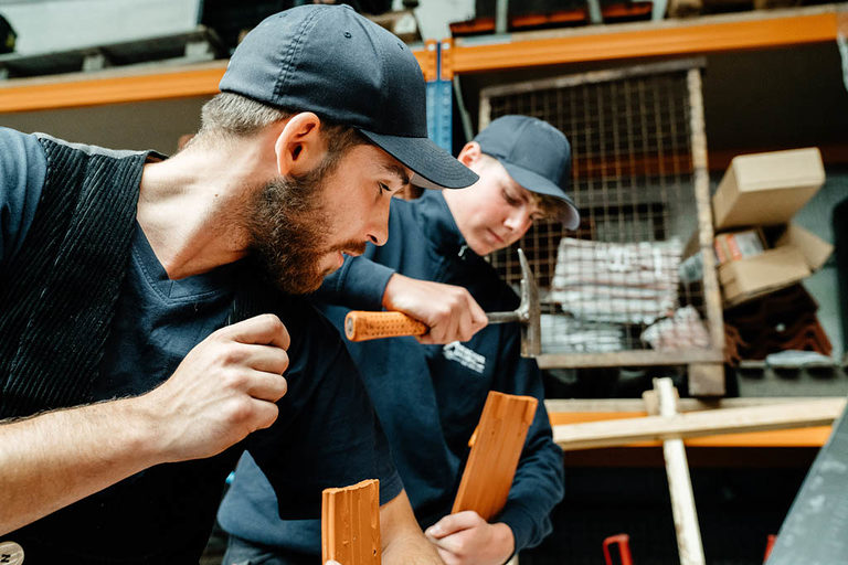 Zwei Dachdecker-Azubis in der Werkstatt, derjenige im Hintergrund mit einem Dachdeckerhammer in der Hand