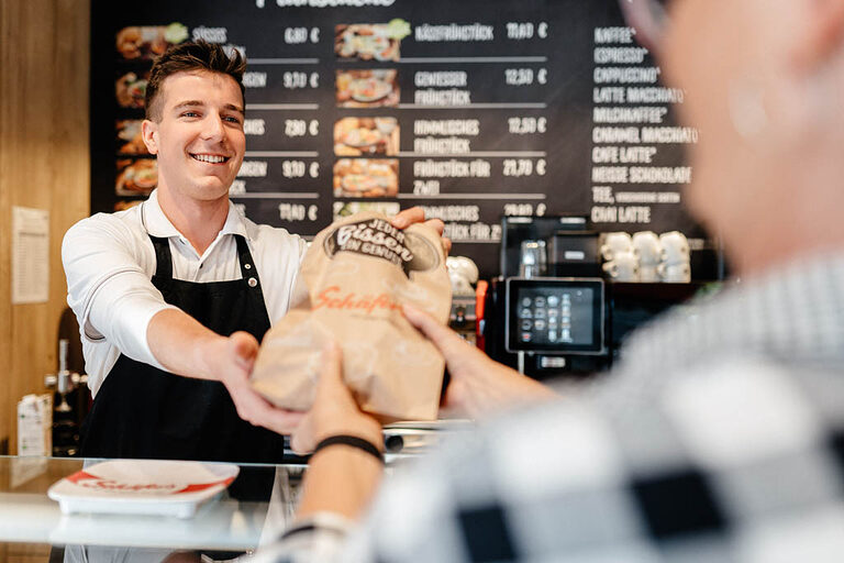 Ein Verkäufer-Azubi reicht in einem Café eine Warentüte an den Kunden