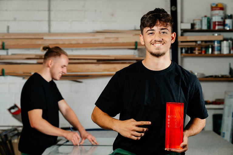 Zwei Glaser-Azubis in der Werkstatt, der vordere hält ein rotes Glasgefäß in der Hand