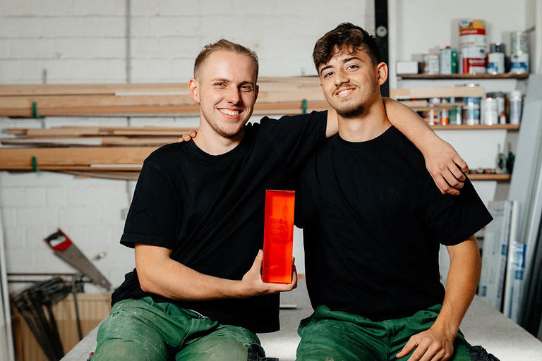 Zwei Glaser-Azubis sitzend in der Werkstatt, der eine hält ein rotes Glasgefäß in der Hand