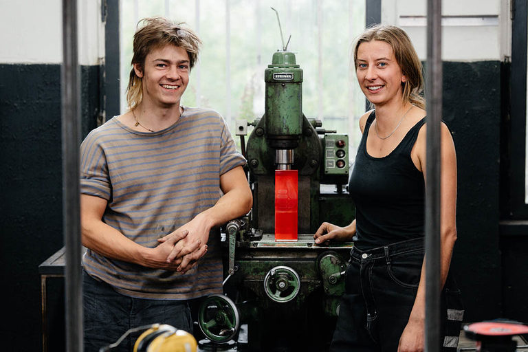 Ein Auszubildender und eine Auszubildende posieren in einer Metallwerkstatt