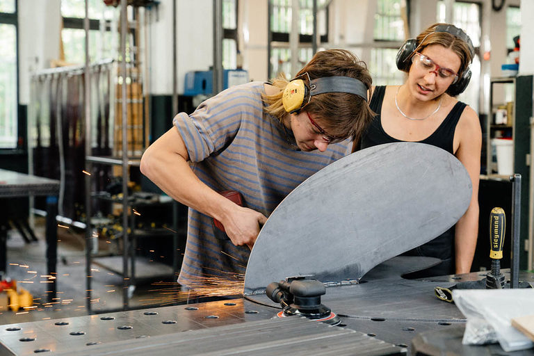 Ein Auszubildender beim Schweißen in einer Metallwerkstatt, eine Auszubildende schaut ihm zu