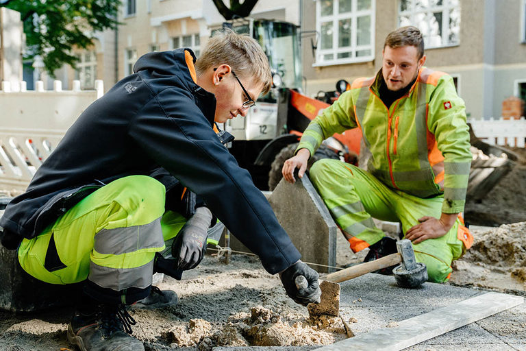 Ein Straßenbauer-Azubi arbeitet mit einem Schonhammer auf der Straße, ein anderer sieht ihm zu.