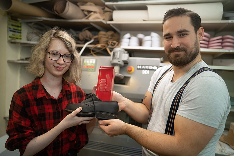 Eine Auszubildende und ein Auszubildender posieren mit einem Schuh in einer orthopädischen Werkstatt.