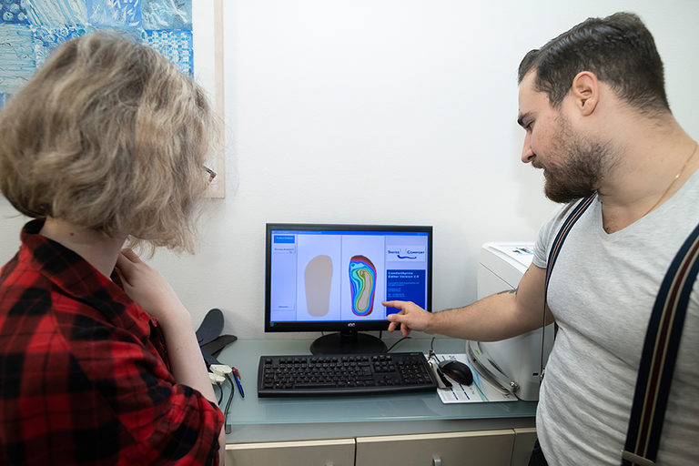 Ein Auszubildender und eine Auszubildende beim Betrachten einer Sohlenabbildung am Monitor in einer orthopädischen Werkstatt.