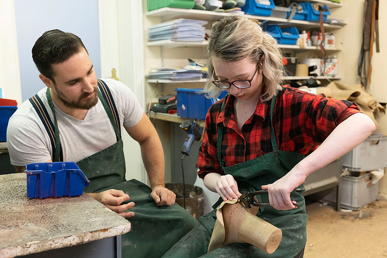 Eine Auszubildende mit einer Schusterzange beim Anfertigen eines Schuhs in einer orthopädischen Werkstatt. Ein Auszubildender schaut ihr bei der Arbeit zu.
