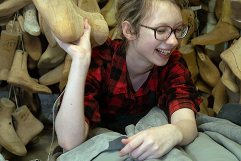 Eine Schuhmacher-Auszubildende mit einem Leisten in der Hand posiert lachend in einer orthopädischen Werkstatt.