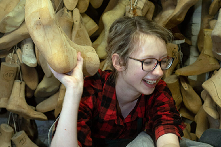 Eine Schuhmacher-Auszubildende mit einem Leisten in der Hand posiert lachend in einer orthopädischen Werkstatt.