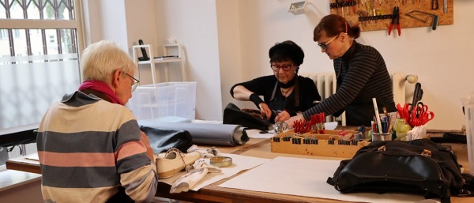 Drei Mitarbeiterinnen der Unikat Taschenmanufaktur &uuml;ben das T&auml;schner Handwerk an einem Tisch aus. 