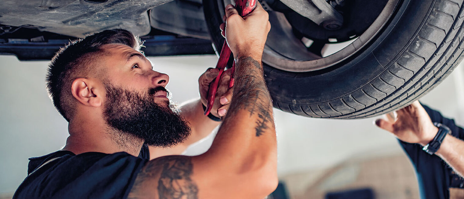 Ein Handwerker steht unter einem Auto in einer KFZ-Werkstatt und repariert das Vorderrad des Autos. 