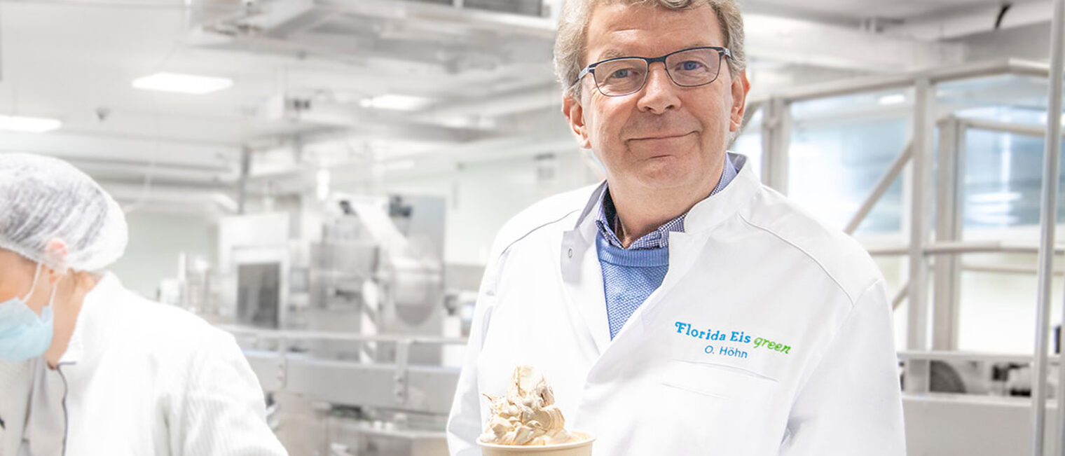 Olaf Höhn in der Produktion von Florida Eis. Olaf Höhn, der Geschäftsführer von Florida Eis, steht in der Produktion von Florida Eis mit einem Becher Eis in der Hand und lächelt in die Kamera.