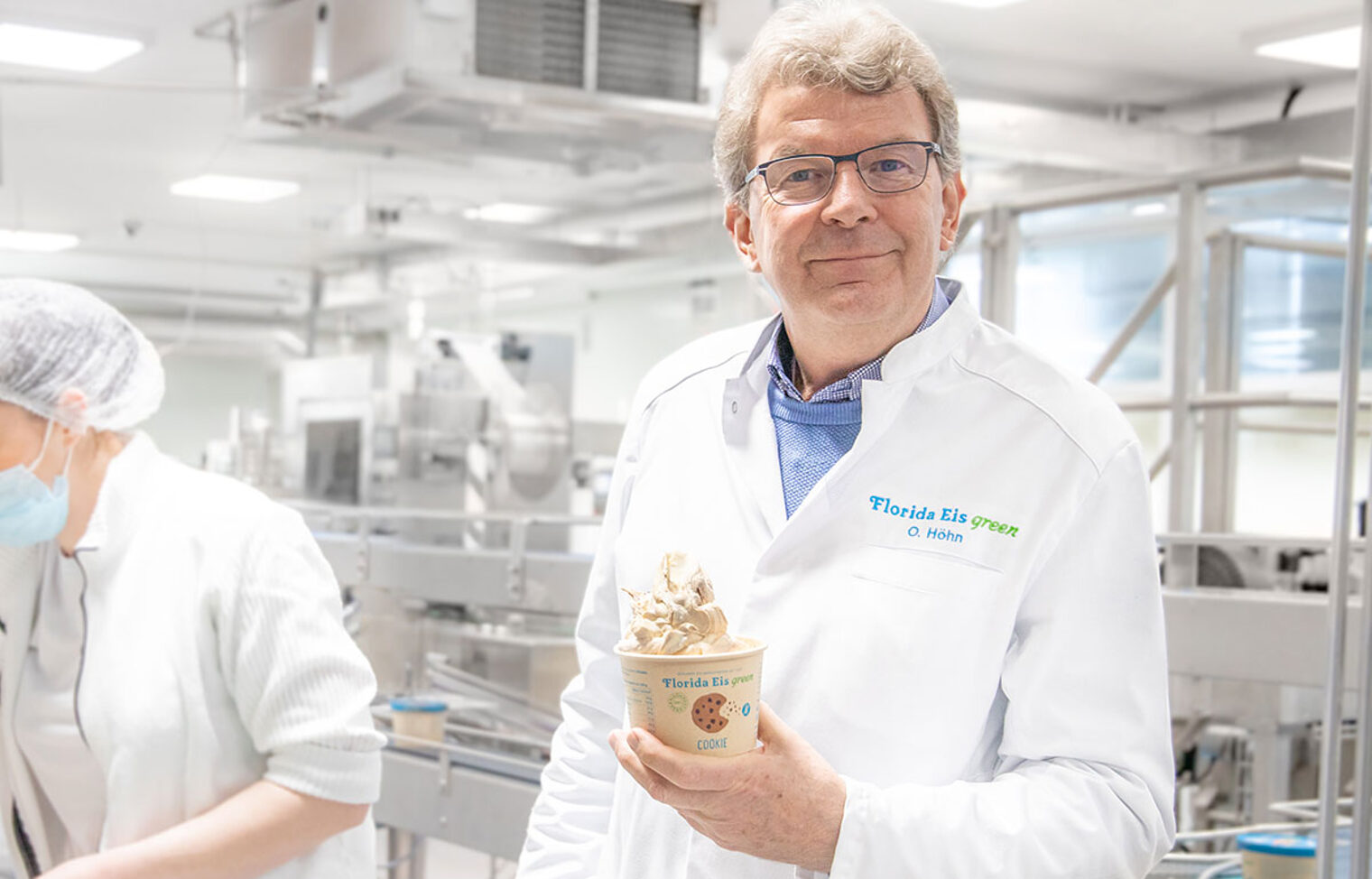 Olaf Höhn, der Geschäftsführer von Florida Eis, steht in der Produktion von Florida Eis mit einem Becher Eis in der Hand und lächelt in die Kamera.