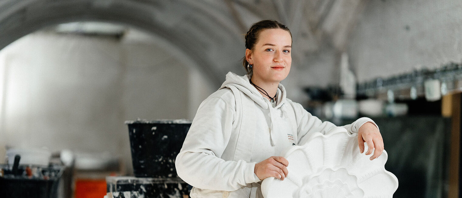 Eine Stuckateurin in weißer Arbeitskleidung hält ein kunstvoll verziertes Stuckelement in Form einer stilisierten Muschel.