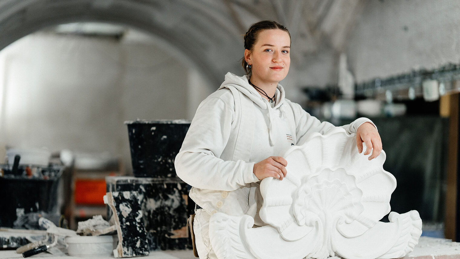 Eine Stuckateurin in weißer Arbeitskleidung hält ein kunstvoll verziertes Stuckelement in Form einer stilisierten Muschel.