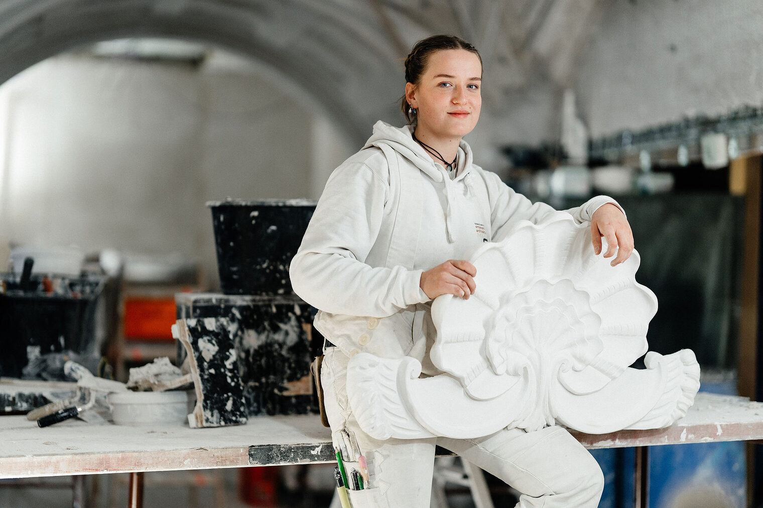 Eine Stuckateurin in weißer Arbeitskleidung hält ein kunstvoll verziertes Stuckelement in Form einer stilisierten Muschel.