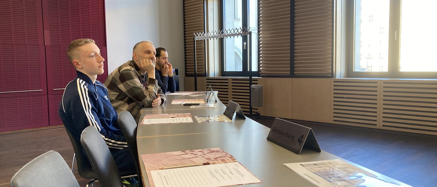 Azubis zu Besuch im Bundesrat: drei Personen sitzen an einem Tisch und hören dem Redner zu.