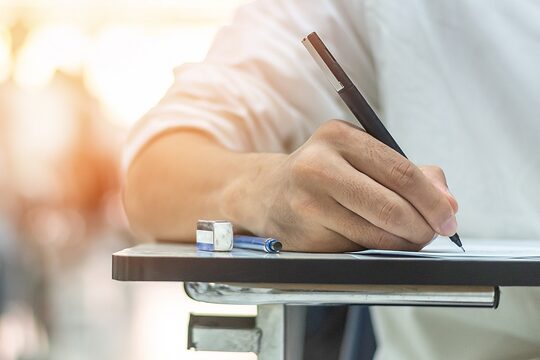 Schulprüfung, Detailaufnahme, Hand mit Stift an einer Unterlage: Schüler/in nimmt an einem Aufnahmetest im Klassenzimmer teil