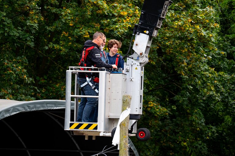 Ein Erwachsener und ein Jugendlicher stehen in einem Baggerkorb vor herbstlich belaubten Bäumen am Tag der Handwerks 2026 im Mellowpark