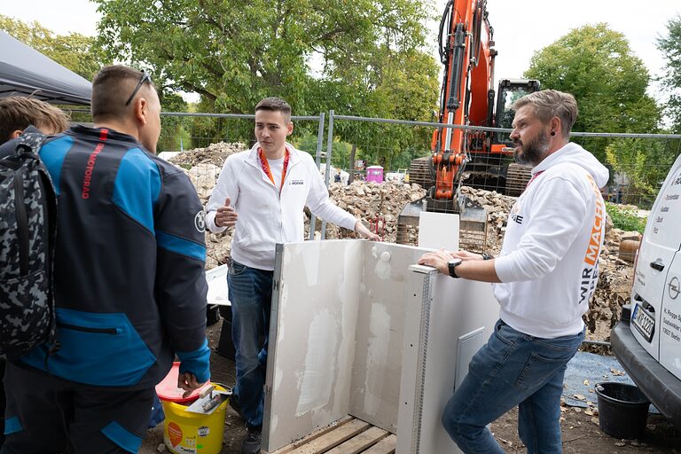 Zwei Standbetreuer im Gespräch mit einem Besucher, im Hintergrund - ein Bagger zu sehe. Tag der Handwerks 2026 im Mellowpark Berlin.