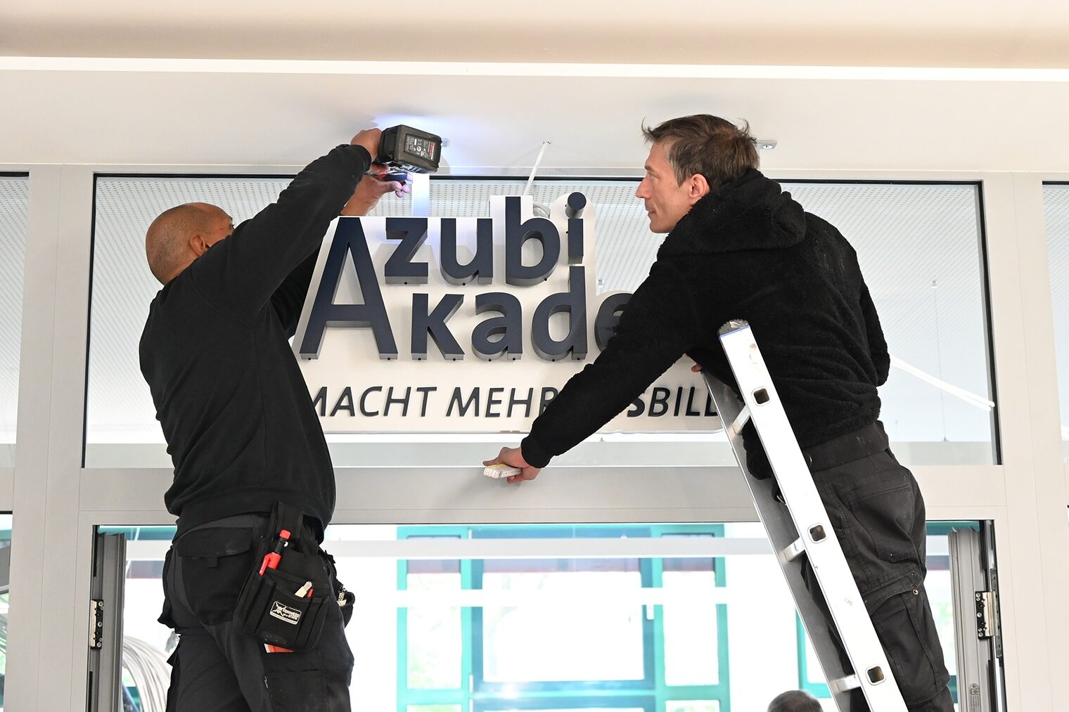 Zwei Bauarbeiter auf einer Leiter: Anbringung des neuen Schilds über dem Eingangsbereich der Azubi Akademie