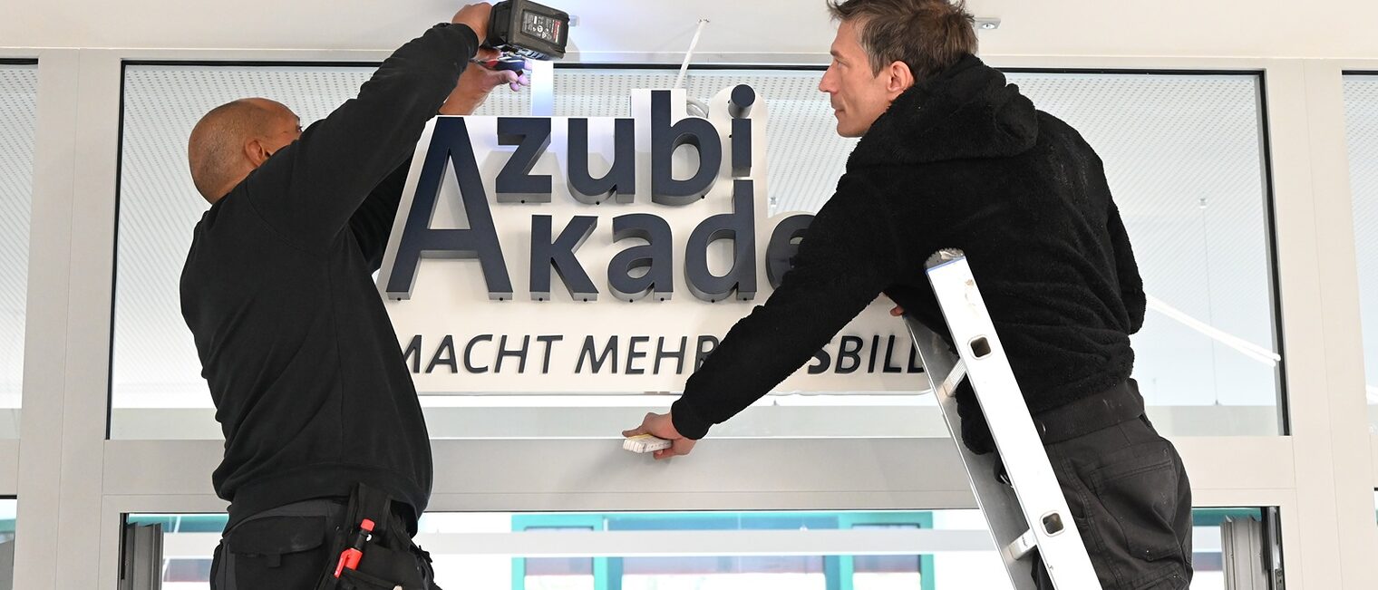 Zwei Bauarbeiter auf einer Leiter: Anbringung des neuen Schilds über dem Eingangsbereich der Azubi Akademie