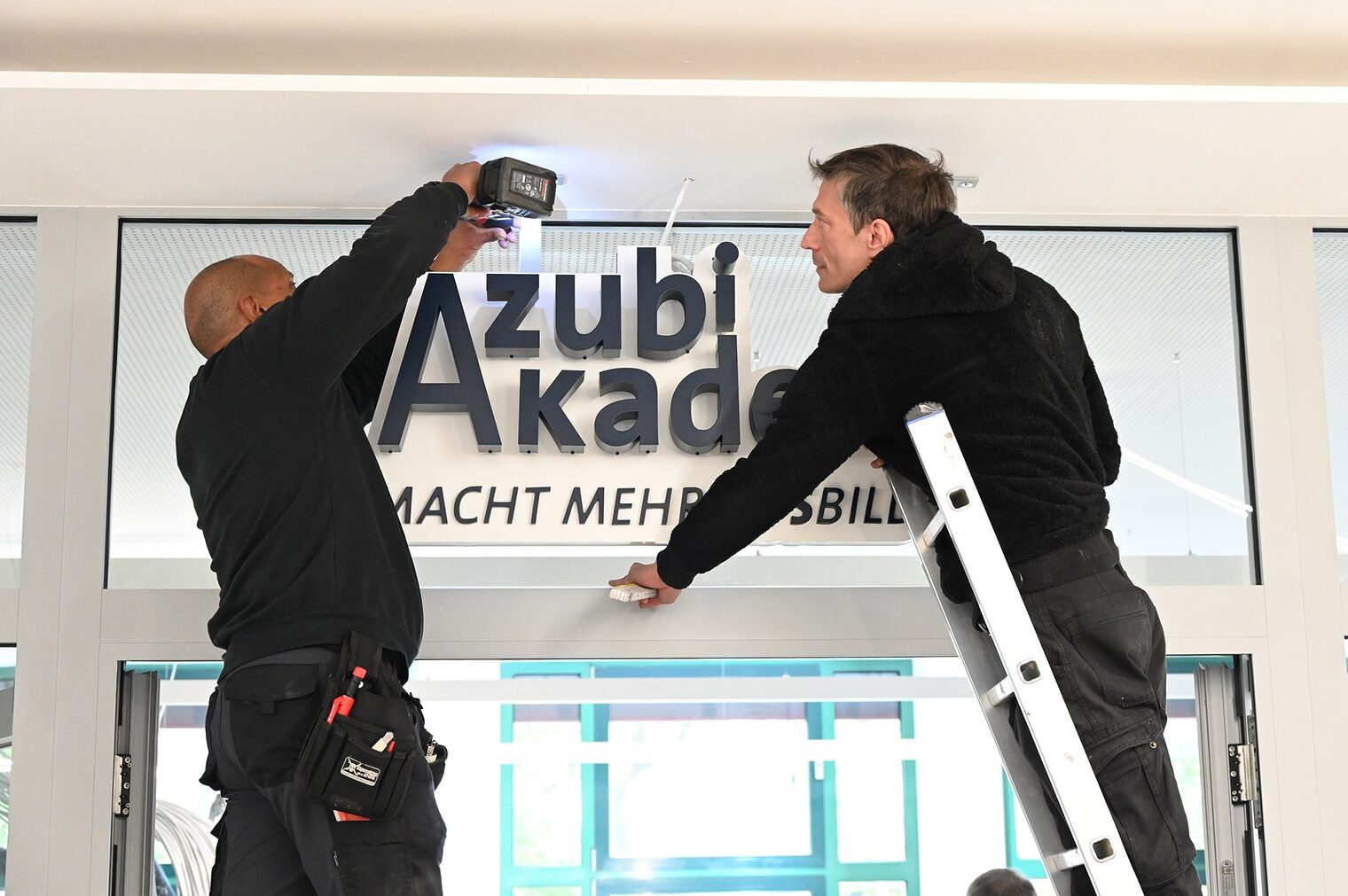 Zwei Bauarbeiter auf einer Leiter: Anbringung des neuen Schilds über dem Eingangsbereich der Azubi Akademie