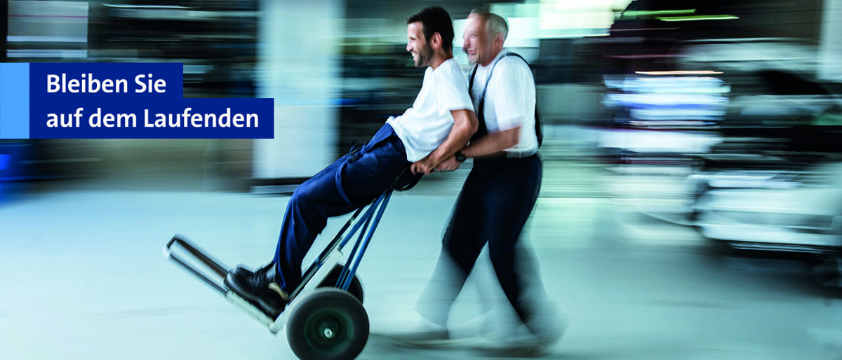 Bleiben Sie auf dem Laufenden mit dem Newsletter der Handwerkskammer Berlin. Zwei Handwerker, die auf einer Sackkarre durch eine Werkstatt fahren.